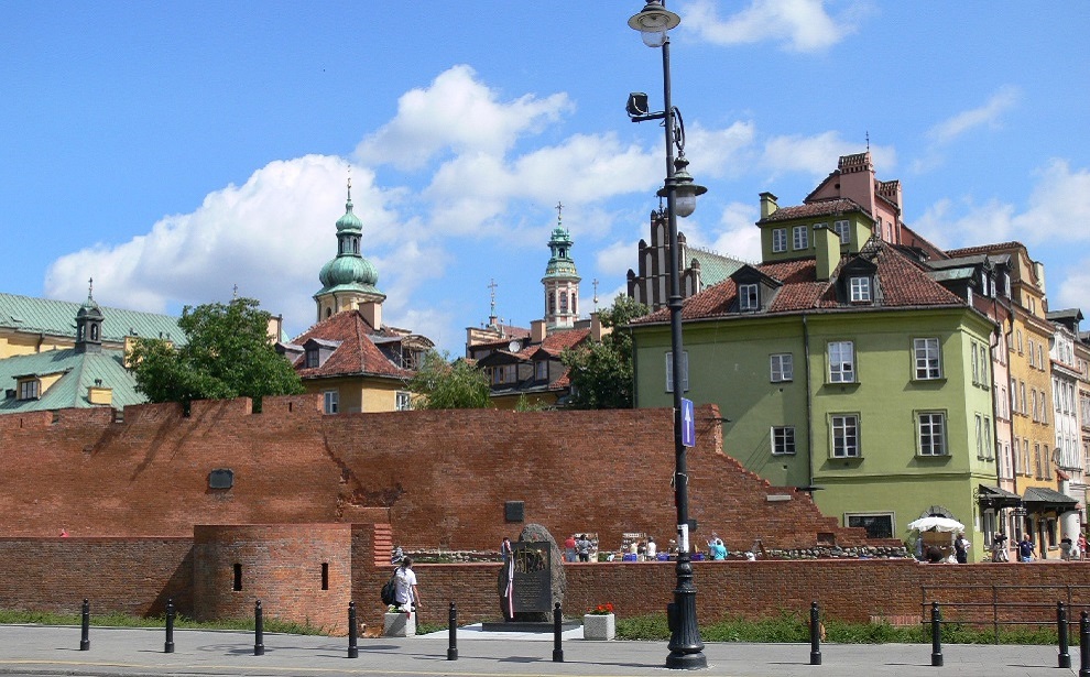 Powiększ obraz: Zdjęcie przedstawia Stare Miasto w Warszawie.  Na pierwszym planie widać ceglane mury obronne, a za nimi kamieniczki i wieże kościołów.
